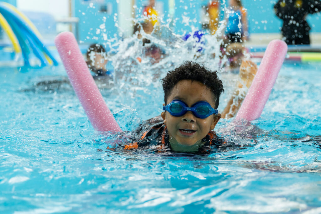Photo of young boy swimming in pool with noodle as swim teacher guides with instruction